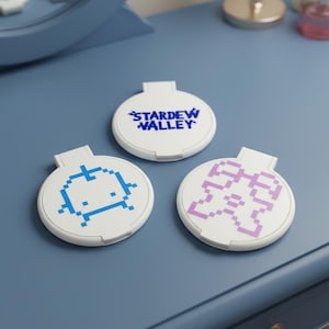 May include: Three white, round compact mirrors with a handle, each featuring a different pixelated design. One mirror has the text "STARDEW VALLEY" in blue, while the others have blue and purple pixel art. The mirrors are on a blue surface.