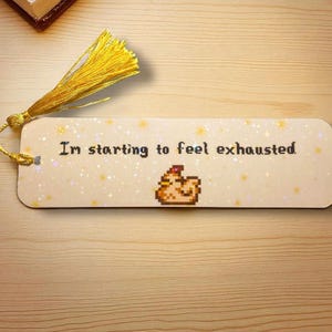 May include: A rectangular bookmark with a light cream background featuring a subtle holographic star pattern. The text "I'm starting to feel exhausted." is printed in black pixelated font above a small pixelated chicken. A yellow tassel is attached.