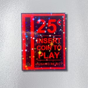 May include: A rectangular magnet with a black background and a red border. The text "25¢" is in large red numbers. The words "INSERT COIN TO PLAY" are in gold lettering. The words "PUSH TO REJECT" are at the bottom.