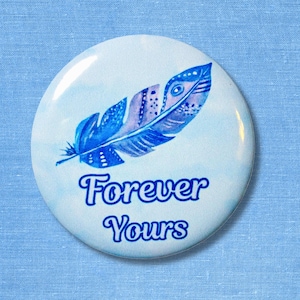 Pode incluir: Um botão redondo com um desenho de pena azul e as palavras "Forever Yours" em fonte cursiva. A pena apresenta um gradiente de tons de azul sobre um fundo azul claro aquarelado. O botão está sobre um tecido azul claro.