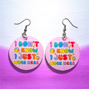 May include: A pair of round, light pink dangle earrings featuring silver-toned metal hooks. The surface of each earring displays the colorful text I DON'T KNOW I JUST WORK HERE in a playful, multi-colored font, accented by small flower icons.