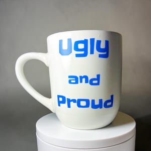 May include: White ceramic mug with the words "Ugly and Proud" in blue block letters. The mug has a curved handle and sits on a white, round surface. The background is a neutral grey.
