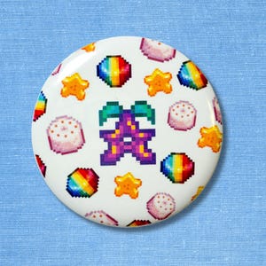 Pode incluir: Um botão branco com um design pixelizado com uma estrela roxa e verde, orbes de arco-íris, estrelas amarelas e sobremesas rosa pixelizadas. O botão é redondo e tem um acabamento brilhante.