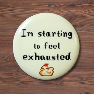 May include: A round, off-white button with the text "I'm starting to feel exhausted" in black pixelated font. A small, pixelated chicken graphic is at the bottom. The button is on a brown wooden surface.