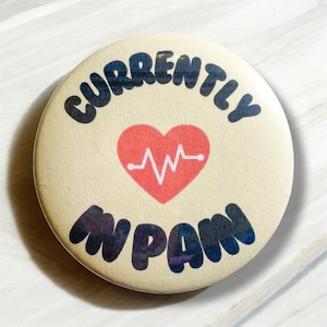 Puede incluir: Un botón redondo con fondo beige y texto negro que dice "Currently in Pain". Un corazón rojo con una línea ECG blanca está en el centro del botón.