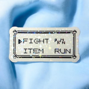 May include: A rectangular sticker with a pixelated design and the words "FIGHT", "ITEM", and "RUN". The sticker has a holographic, glittery effect with a white border and is set against a light blue background.