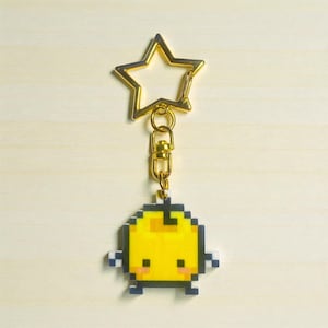 Könnte beinhalten: Ein goldfarbener sternförmiger Schlüsselanhänger mit einem pixeligen gelben Charakter. Der Charakter hat kleine schwarze Augen und Arme und ist mit einem goldfarbenen Verschluss an dem Stern befestigt. Der Hintergrund ist ein helles Holzmaserung.