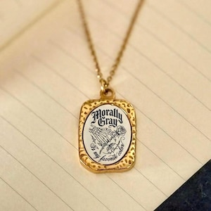 May include: Gold-coloured necklace with a rectangular pendant. The pendant displays the text "Morally Gray" above a graphic of praying hands and a rose. The words "If my favourite colour" are also visible. The necklace rests on a notebook.