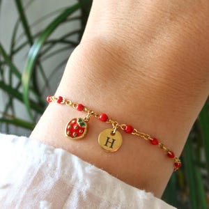 Op de afbeelding: Gouden armband met rode kralen en bedels. Een rode aardbeibedel met groene blaadjes en een gouden schijf met de letter "H" zijn zichtbaar. De armband wordt om een pols gedragen.