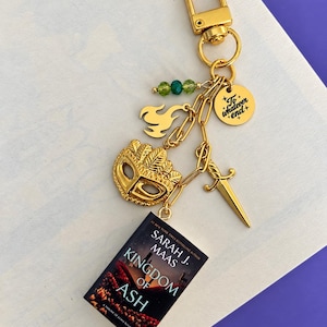 Könnte beinhalten: Ein goldfarbener Metallschlüsselanhänger mit einer Miniaturbuch-Nachbildung von Kingdom of Ash von Sarah J. Maas, einem Maskenanhänger, einem Schwert, einer Flamme, drei grünen Perlen und einer Scheibe mit der Gravur To whatever end.