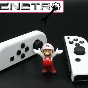 Pode incluir: Dois controladores brancos Nintendo Switch Joy-Con com botões pretos e um stick analógico preto. Os controladores estão em uma superfície preta com uma pequena figura do Mario no meio.
