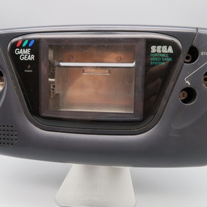 Könnte beinhalten: Eine schwarze Sega Game Gear Handheld-Videospielkonsole mit einem transparenten Kunststoffbildschirm. Die Konsole hat einen Power-Knopf, einen Start-Knopf und einen Kartensteckplatz. Der Text "GAME GEAR" und "SEGA PORTABLE VIDEO GAME SYSTEM" ist auf der Konsole aufgedruckt.