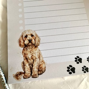 May include: A lined notepad featuring a watercolour illustration of a light brown dog sitting with its tail curled behind it. The dog is surrounded by a light brown watercolour background. The bottom of the notepad has four black paw prints.