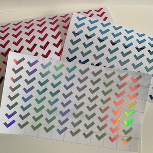 May include: Sheets of stickers featuring checkmark designs in various colours. The stickers include red, blue, and iridescent options. The checkmarks are arranged in a grid pattern on a white background.