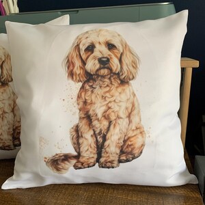 May include: White square throw pillow featuring a watercolor illustration of a golden doodle dog. The dog is sitting, with a soft, fluffy coat and expressive eyes. Brown watercolor splatters surround the dog, adding a touch of artistic flair. The pillow is on a wooden surface.