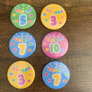 May include: Six colourful round buttons with numbers 3, 5, 7, and 10. Each button has a different colour background, including green, yellow, blue, and pink. The numbers are in white, with star and banner decorations.