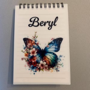 May include: A spiral-bound notepad with the name "Beryl" in elegant script. The notepad features a colorful butterfly design with floral accents in shades of blue, orange, and pink. The background has lined paper.
