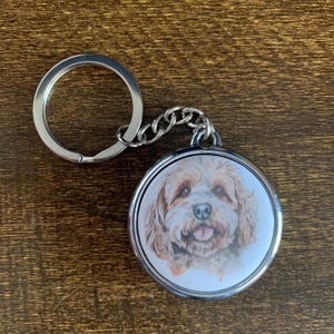 May include: A round, silver-toned keychain featuring a watercolor-style portrait of a light brown and white dog with a happy expression. The dog's tongue is visible. The keychain has a split ring and a chain link attachment.