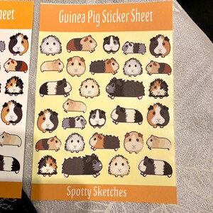 May include: A sheet of stickers featuring various cartoon guinea pigs in different poses and colours. The sheet is yellow with the text "Guinea Pig Sticker Sheet" and "Spotty Sketches" printed on it.