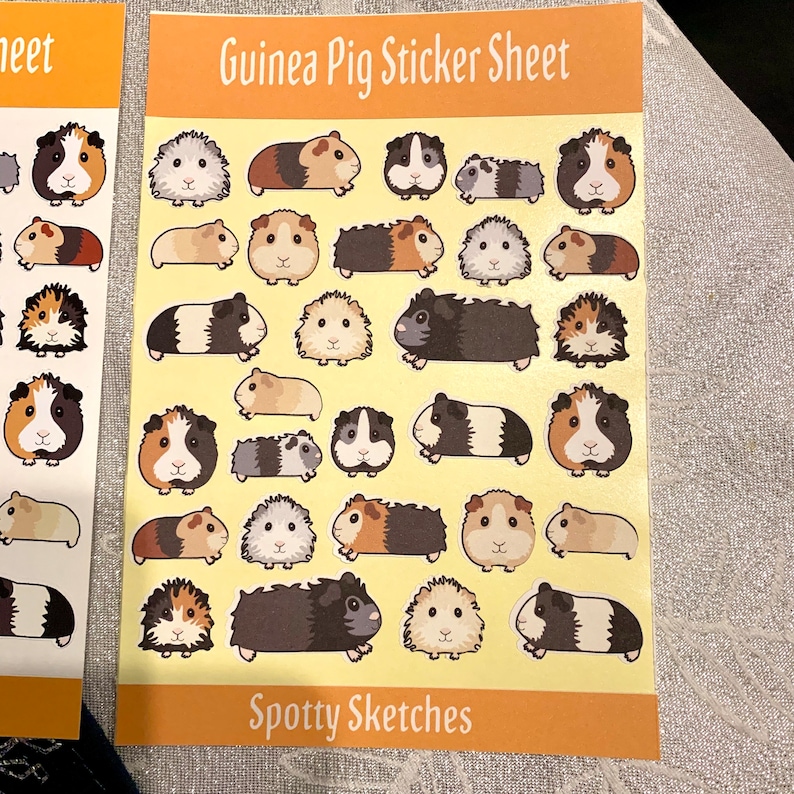 May include: A sheet of stickers featuring various cartoon guinea pigs in different poses and colours. The sheet is yellow with the text "Guinea Pig Sticker Sheet" and "Spotty Sketches" printed on it.