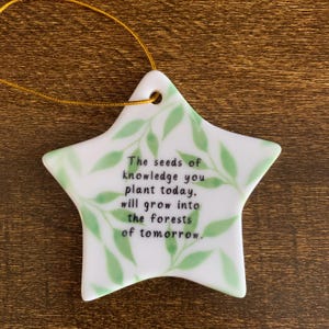 May include: A star-shaped ceramic ornament with a gold string for hanging. The ornament is white with green leaf designs and the text: "The seeds of knowledge you plant today, will grow into the forests of tomorrow."