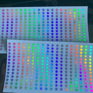 Op de afbeelding: Twee vellen wit papier met rijen van regenboogkleurige, holografische ster stickers. De sterren zijn gerangschikt in regenboogkleuren.