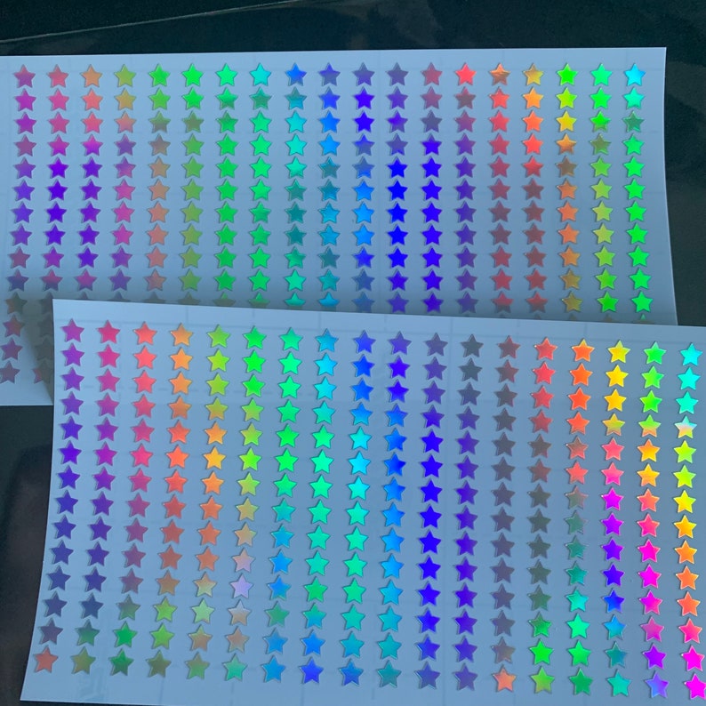 Op de afbeelding: Twee vellen wit papier met rijen van regenboogkleurige, holografische ster stickers. De sterren zijn gerangschikt in regenboogkleuren.