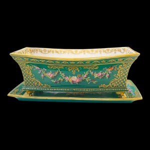 Puede incluir: Una jardinera rectangular de porcelana verde esmeralda con una base rectangular a juego. La jardinera presenta adornos dorados y diseños florales y de volutas. La base tiene pequeños pies. La jardinera es probablemente para uso decorativo.