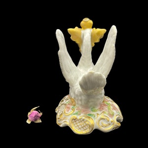 May include: A white porcelain swan figurine with gold accents and a yellow flower on its head. The swan is standing on a base with floral designs and gold accents.