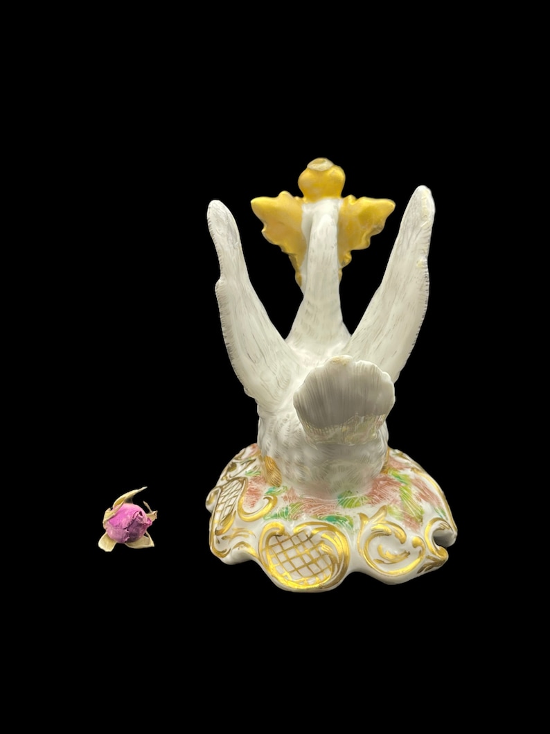 May include: A white porcelain swan figurine with gold accents and a yellow flower on its head. The swan is standing on a base with floral designs and gold accents.