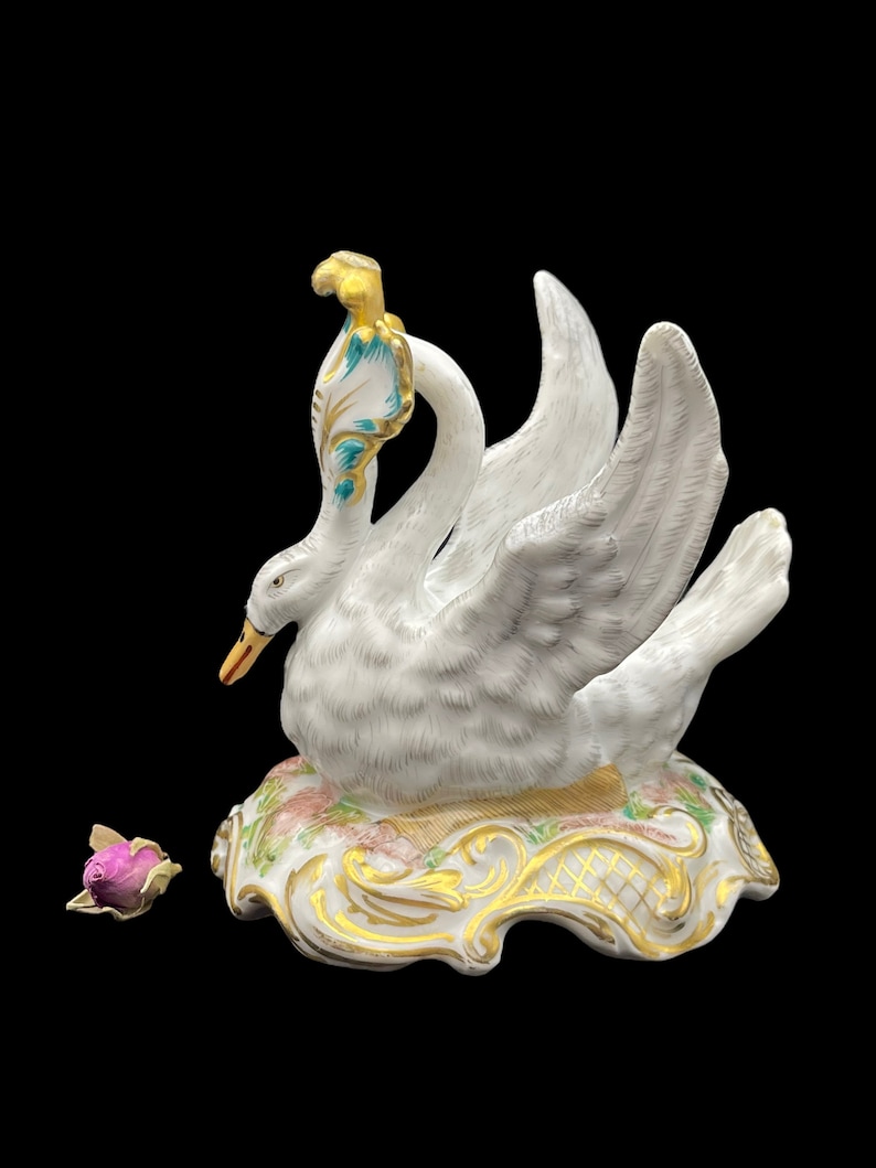May include: A white porcelain swan figurine with gold and blue accents. The swan is perched on a base with floral details and gold trim. A small pink rose is in the foreground.