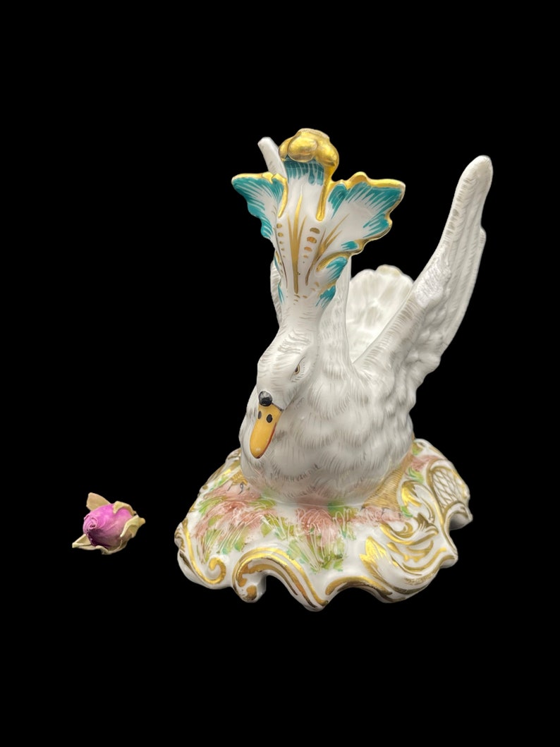 May include: A white porcelain swan figurine with gold and teal accents. The swan is perched on a base with floral details and gold trim.