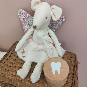 Puede incluir: Un ratón de peluche blanco con alas florales rosas y un tutú rosa, sentado en una cesta de mimbre con una caja de madera en forma de diente.