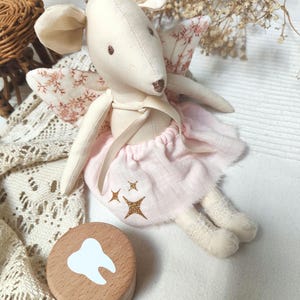 Op de afbeelding: Een zachte, crèmekleurige muizenpop met grote oren, gekleed in een roze tutu en bloemen vleugels. Een houten tandendoos met een witte tandafbeelding staat in de buurt. De pop heeft een grillig ontwerp.