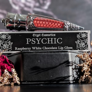 May include: A silver and red lip gloss tube rests on a black box with the text "PSYCHIC Raspberry White Chocolate Lip Gloss." The box is decorated with white rose illustrations. The background is a blurred pattern with red roses and gold accents.