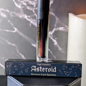 May include: A black tube of Crypt Cosmetics Asteroid Duochrome Liquid Eyeshadow. The tube has a gold label with the product name and a constellation design.