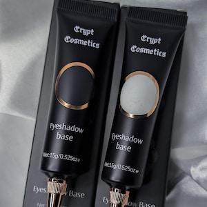 Peut inclure: Deux tubes noirs de base pour fard à paupières Crypt Cosmetics, chacun avec un accent doré et un embout doré. Les tubes sont étiquetés "Eyeshadow base" et "Net: 15g/0.525oz". Le fond est un tissu clair et soyeux.
