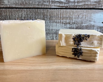 Puede incluir: Dos barras de jabón hechas a mano, una lisa y otra con flores de lavanda secas incrustadas en la parte superior. El jabón es blanco y se encuentra sobre una superficie de madera.