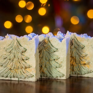Puede incluir: Tres pastillas de jabón rectangulares con un diseño de árbol de Navidad. El jabón es de color verde claro con un árbol detallado en verde más oscuro, acentuado con motas doradas. Los bordes son de color azul claro y el fondo está borroso con luces bokeh.