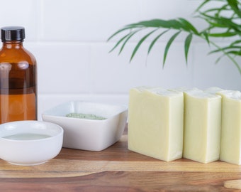 May include: Three bars of light green soap on a wooden surface next to a clear glass bottle with a black lid, a white bowl with green powder, and a white bowl with clear liquid.