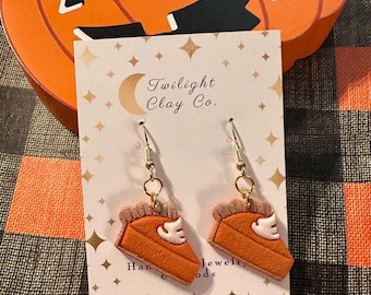 May include: A pair of dangle earrings with a pumpkin pie slice design. The earrings are made of polymer clay and have a gold finish. The earrings are on a white card with the text "Twilight Clay Co."
