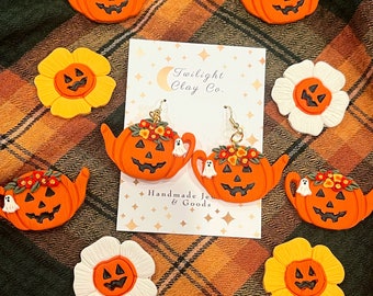 Può includere: Un paio di orecchini fatti a mano in argilla polimerica a forma di teiera a forma di zucca con dettagli floreali e fantasmi. Gli orecchini sono esposti su una carta con il testo "Twilight Clay Co. Handmade Jewelry & Goods".