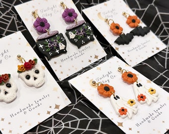 Può includere: Quattro carte con orecchini in argilla polimerica a tema Halloween. Gli orecchini sono esposti su carte bianche con il testo "Twilight Clay Co." e "Gioielli fatti a mano e articoli". Gli orecchini includono un calderone con fantasmi, un teschio con fiori, un pipistrello con fiori arancioni e un fantasma con fiori arancioni.