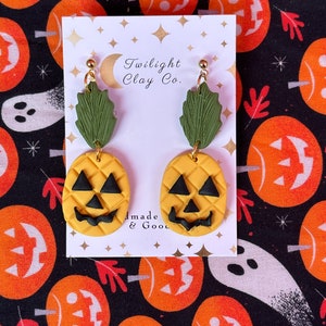 May include: A pair of pumpkin-shaped earrings with green leaves. The earrings are made of yellow and black clay and have a cute, friendly face. The earrings are on a white card with the text "Twilight Clay Co." and "Handmade & Good".