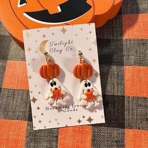 以下が含まれることがあります： 白い幽霊がミニチュアのオレンジ色のカボチャを持っている、手作りのポリマークレイのイヤリングのペア。イヤリングには「Twilight Clay Co. Handmade Jewelry & Goods.」のラベルが付いています。