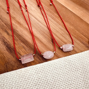May include: Three red string bracelets with pink crystal charms. The charms are a rectangle, an oval, and a square.