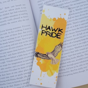 May include: A yellow bookmark with a hawk illustration and the text "HAWK PRIDE" in black lettering. The hawk is flying with its wings spread and is silhouetted against a yellow and orange watercolor background.