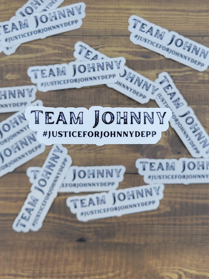 May include: White stickers with the text "TEAM JOHNNY" and "#JUSTICEFORJOHNNYDEPP" in a dark font. The stickers are scattered on a wooden surface. The stickers are rectangular with rounded corners.