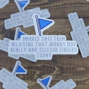 May include: A white sticker with a blue flag design and the text "Marked safe from believing that Johnny Depp really had scissor fingers today".