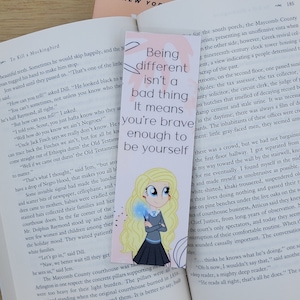 May include: A bookmark featuring a cartoon girl with blonde hair and blue eyes. She is wearing a blue and white school uniform and holding a blue wand. The bookmark has a pink background and the text "Being different isn't a bad thing. It means you're brave enough to be yourself."